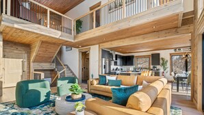 Un'ampia zona giorno con soffitto alto, travi in legno, un balcone e una cucina sullo sfondo.