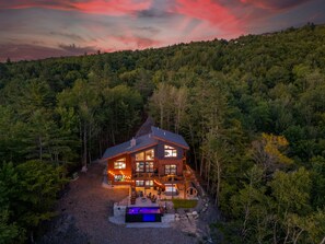 A cabin with a large deck, a hot tub, and a view of a forested hillside.