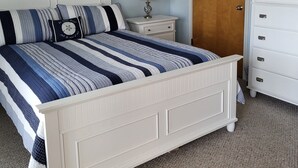 Une chambre à coucher avec un lit blanc, une table de chevet avec une lampe, une commode, un ventilateur de plafond et une porte.