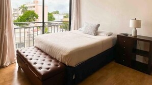 A bedroom with a large bed, a bench, a nightstand, and a view of the city through a large window.