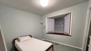 A bedroom with a single bed, a window with blinds, a floor lamp, and a carpeted floor.
