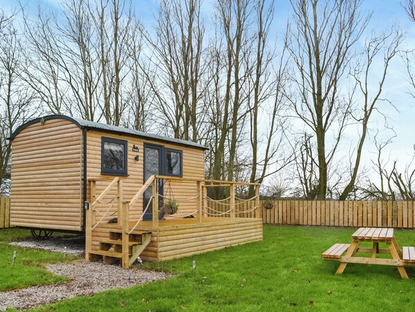 A wooden cabin with a staircase leading to the entrance, a small deck, and a picnic table in a grassy area with trees in the background.
