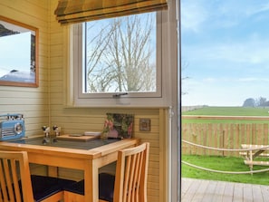 A wooden interior with a table, chairs, a radio, a framed picture, and a window with a view of a fence and greenery.