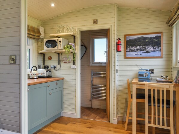A compact kitchen with a microwave, toaster, and kettle on the counter, a small table with a radio, and a framed picture on the wall.