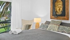 A bedroom with a bed, a ceiling fan, a Buddha statue, and a view of greenery.