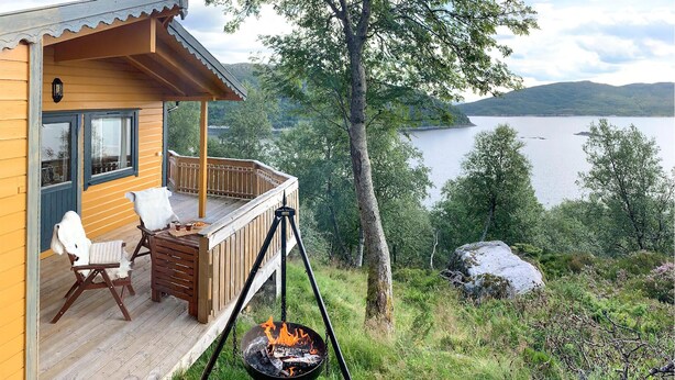 A wooden cabin with a deck, a hanging fire pit, and a view of a lake and mountains.