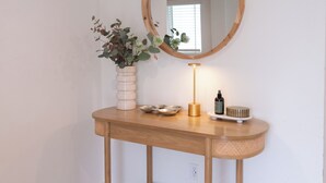 A wooden console table with a round mirror, a vase with green plants, a lamp, and a small shelf with a bottle and a bowl. - Central Thousand Oaks retreat close to shops & eats! (Thousand Oaks)