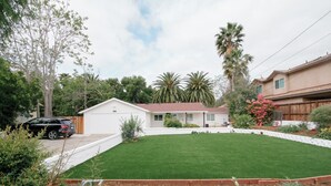 A residential property with a well-maintained lawn, a small house with a red roof, and surrounding greenery including palm trees and flowering plants. - Central Thousand Oaks retreat close to shops & eats! (Thousand Oaks)