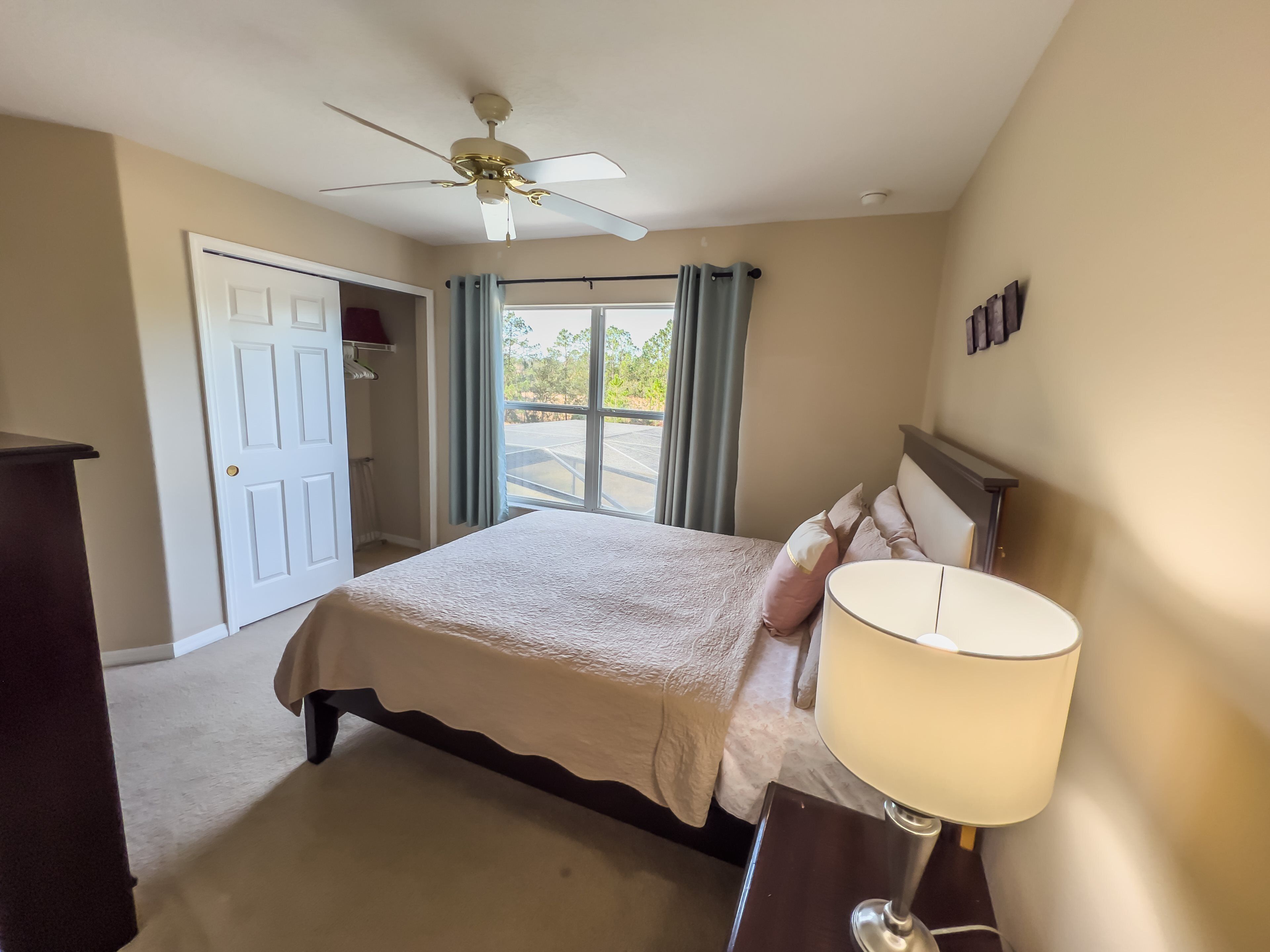 Une chambre à coucher avec un lit, un ventilateur de plafond, une table de chevet avec une lampe, et une vue sur l’extérieur par une fenêtre.