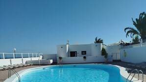 A clear blue pool with a white poolside area, a white building with a door and windows, and a palm tree in the background. - Dream getaway! seaview shell (Playa Honda , lanzarote)