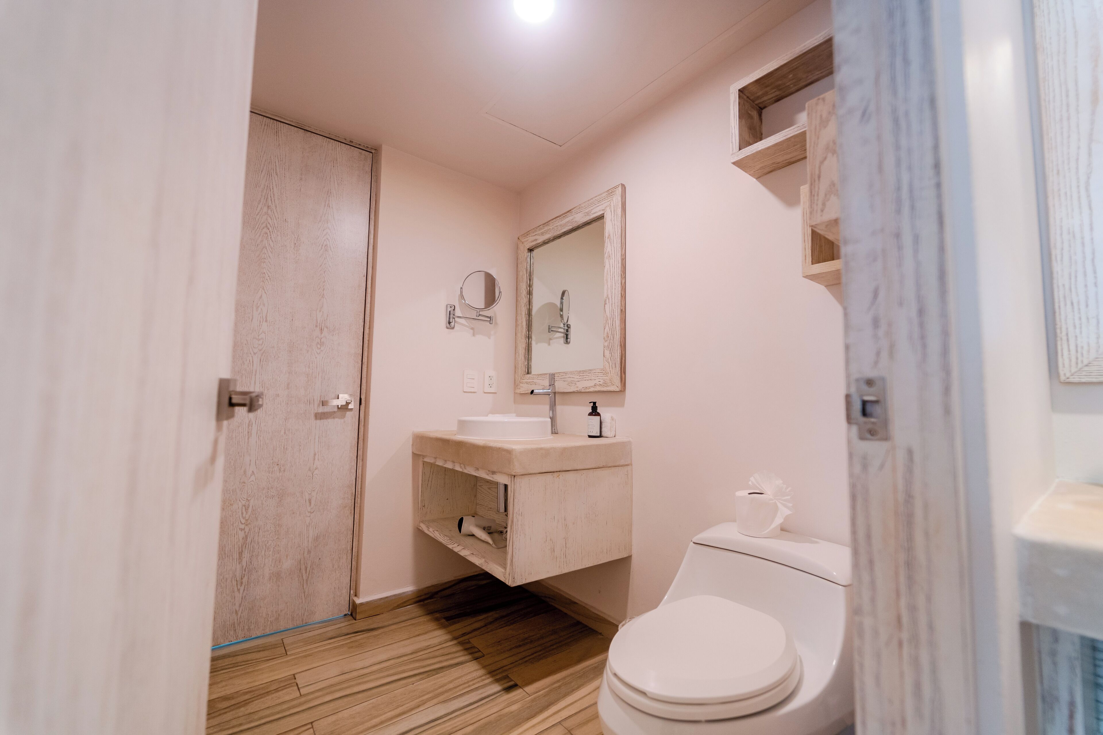 A bathroom with a wooden floor, a white toilet, a sink with a mirror above it, and a shelf with toiletries.