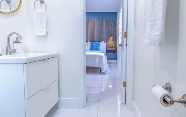 A modern bathroom with a white sink, toilet, and a door leading to a bedroom with a bed and a nightstand.