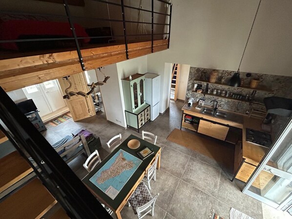 Une chambre de style loft avec un lit superposé, un coin repas agrémenté d’une carte, une cuisine avec un mur arrière en pierre, et une vue sur l’extérieur.