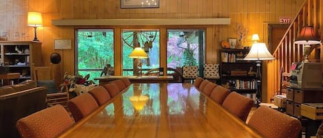 Een ruime houten eettafel omringd door stoelen met kussens in een kamer met houten wanden en grote ramen.
