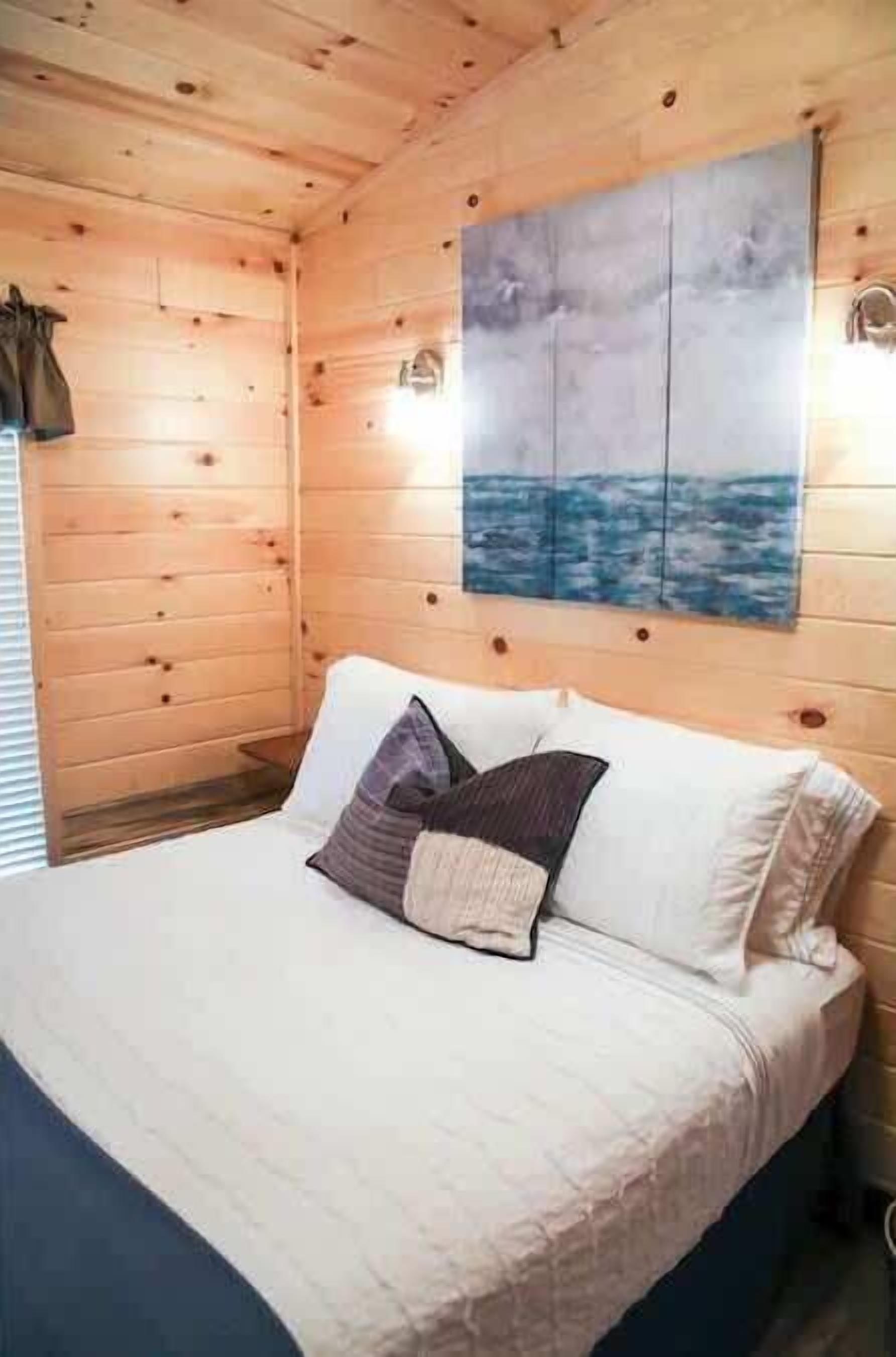 A wooden cabin room with a bed, a painting of a seascape, and a window with blinds.