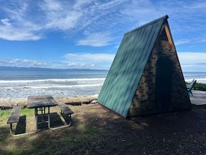 Una estructura de madera con techo inclinado cerca de la playa, una mesa de picnic y vistas al océano.