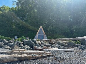 Une petite cabane dotée d’un toit triangulaire, entourée de gros rochers et de morceaux de bois flotté, se dressant sur un fond de forêt dense.