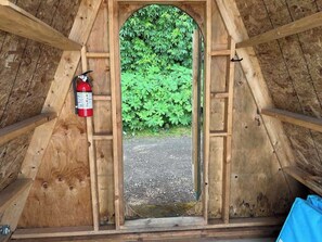 A wooden attic room with a fire extinguisher mounted on the wall and a view of a gravel path leading to a green bush.