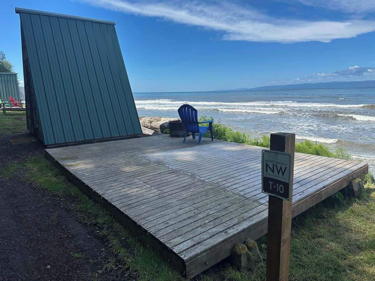 Une terrasse en bois avec une chaise bleue et une structure verte, donnant sur une plage où l’on peut voir les vagues.