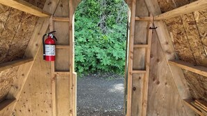 A wooden attic room with a fire extinguisher mounted on the wall, a window with a view of green foliage, and a gravel floor. - Whiskey Creek Beach Front A-Frame Site 11 (Port Angeles)