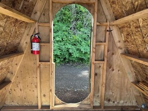 Um quarto de sótão com piso de cascalho, janela com vista para vegetação verde e um extintor de incêndio fixado na parede.