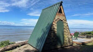 A small, triangular wooden structure with a green roof, located near a body of water. - Whiskey Creek Beach Front A-Frame Site 11 (Port Angeles)