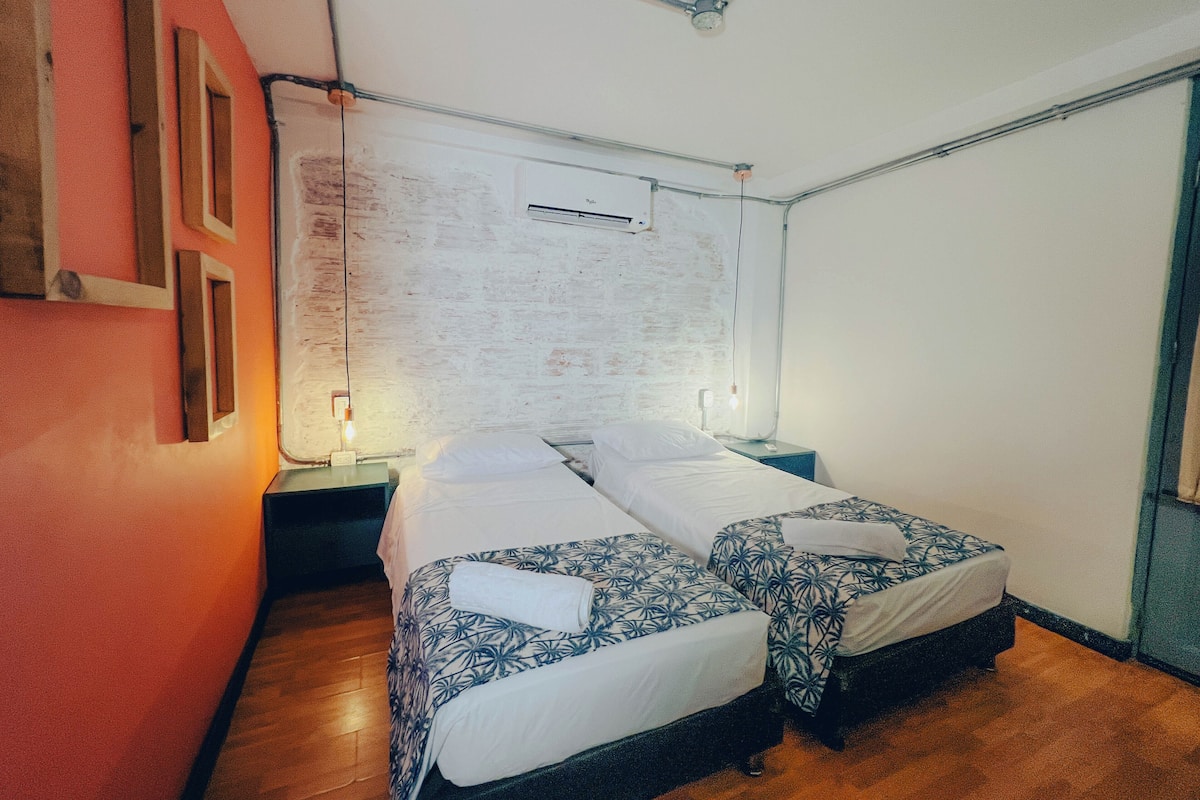 Une chambre avec deux lits, un plancher en bois, un mur blanc orné d’un tableau d’affichage et un ventilateur de plafond.