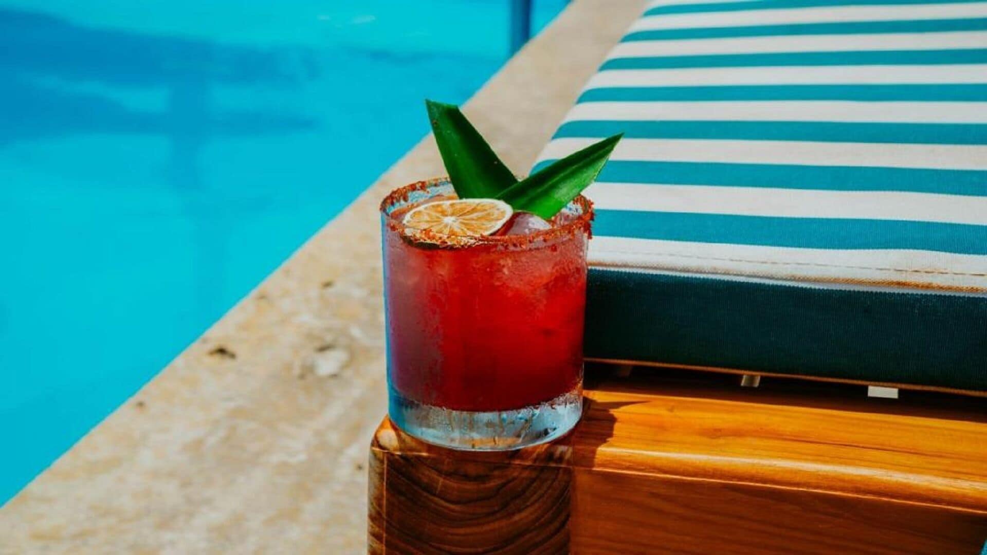 A cocktail with a garnish on a wooden coaster by a poolside lounge area.