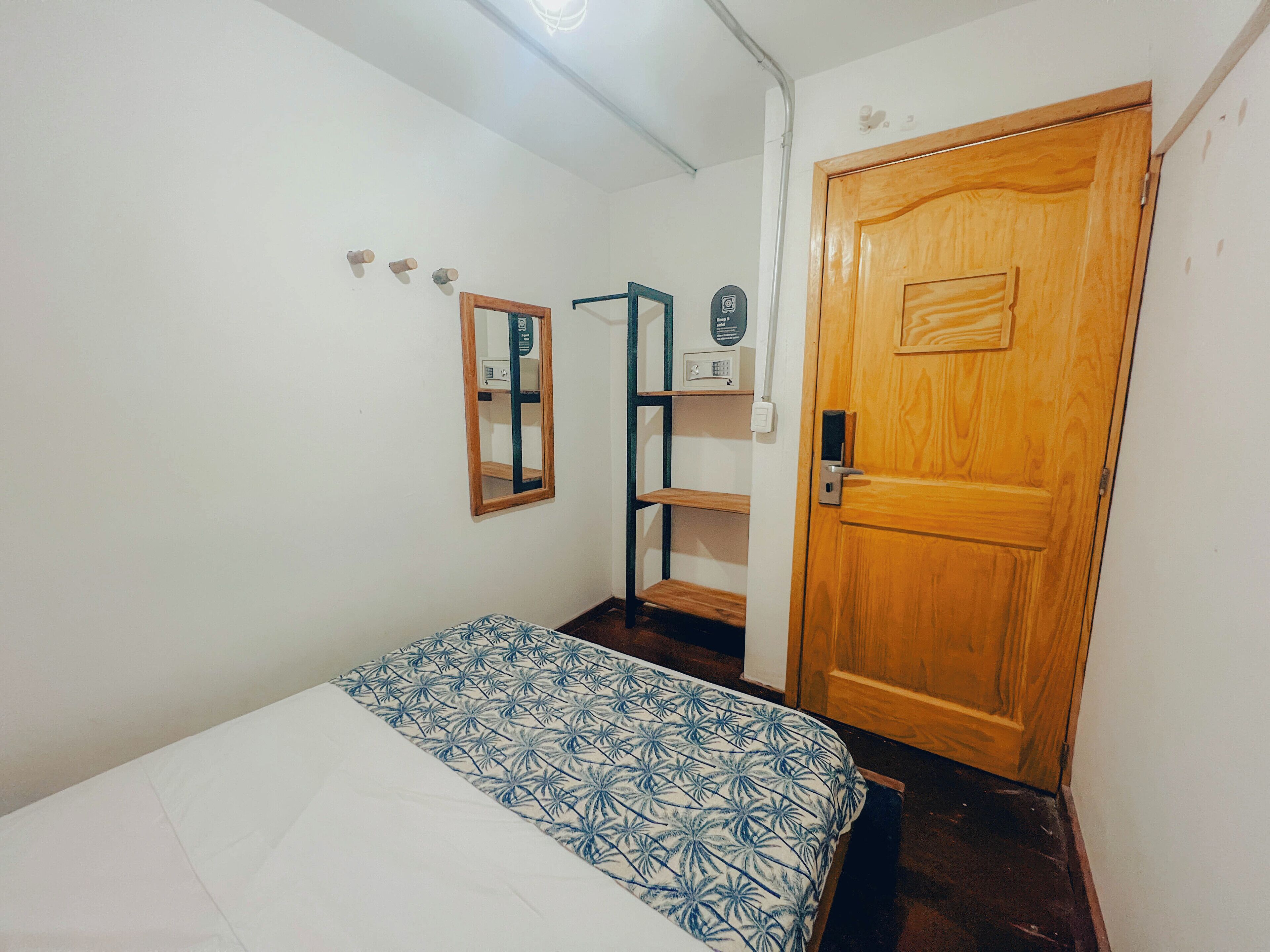 A small, sparsely furnished room with a bed, a wooden door, a mirror, and a shelf unit.
