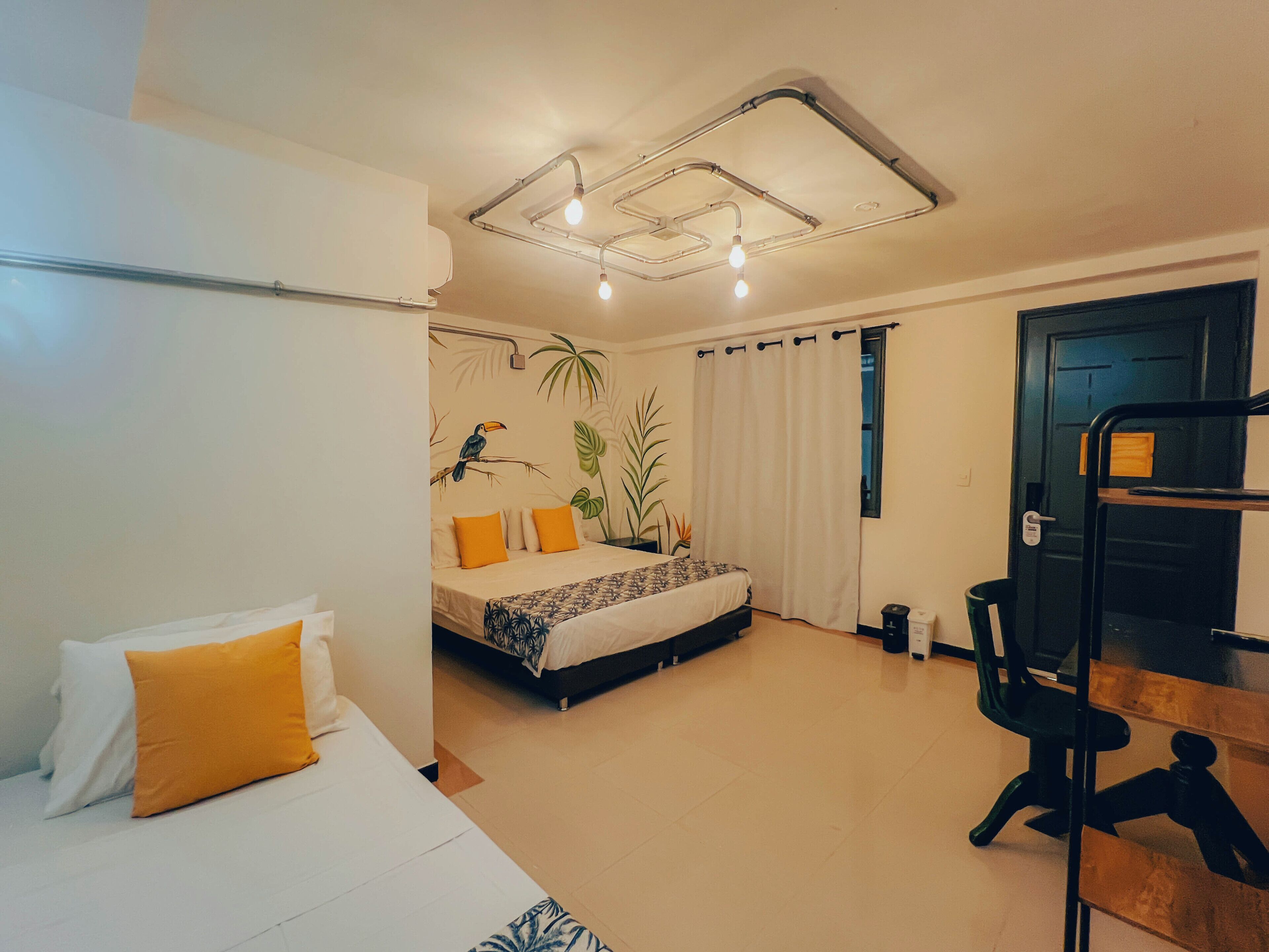 A bedroom with a bed, a desk, and a chair. There is a window with curtains, a door, and a ceiling with exposed pipes and lights.