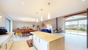 Una cocina moderna con una isla central, un comedor con una mesa grande y sillas, y vistas al exterior a través de amplios ventanales.