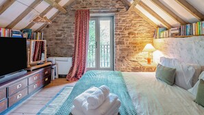 A bedroom with a stone wall, wooden beams, a bed, a TV, bookshelves, and a window with curtains.