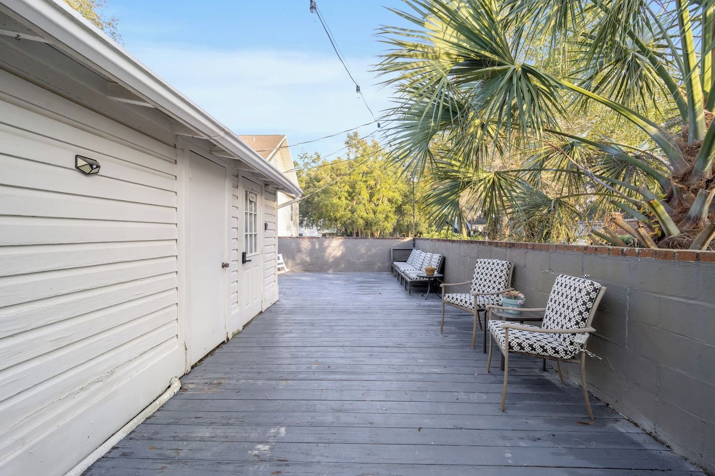 A covered outdoor deck with wooden flooring, two patterned chairs, and a palm tree.
