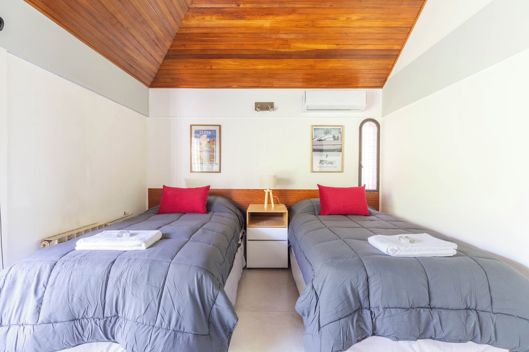 A room with two beds, each with a red pillow and a white towel. There is a wooden ceiling and a small table between the beds.