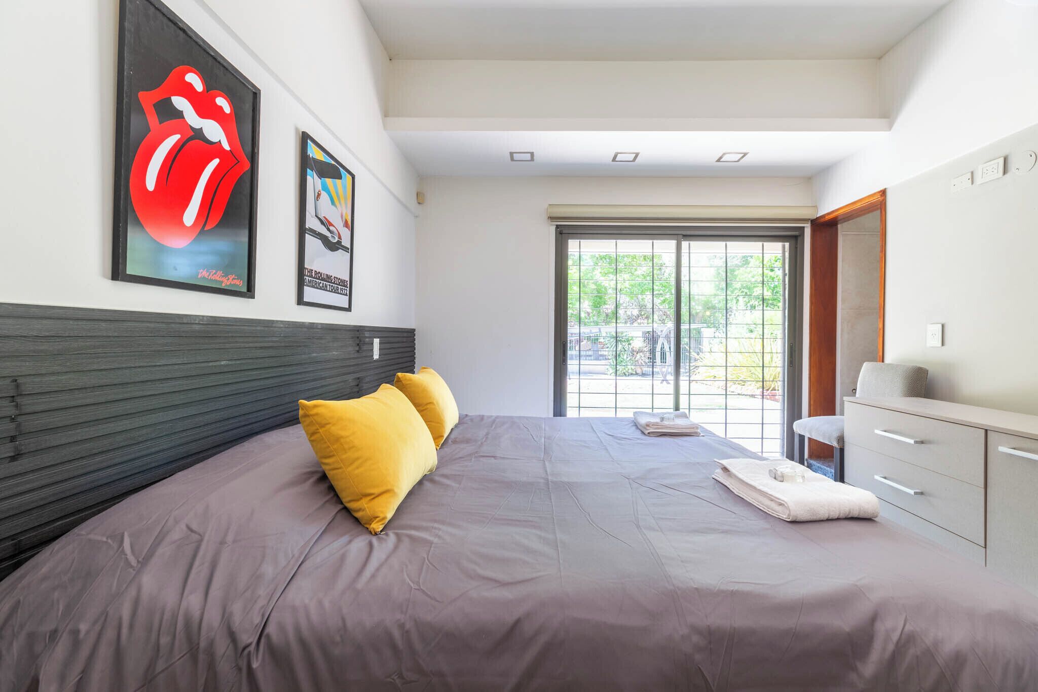 Een moderne slaapkamer met een groot bed, twee gele kussens, een poster van de Rolling Stones in een lijst, een badkamer met een wit wastafelmeubel, en uitzicht op groen door het raam.