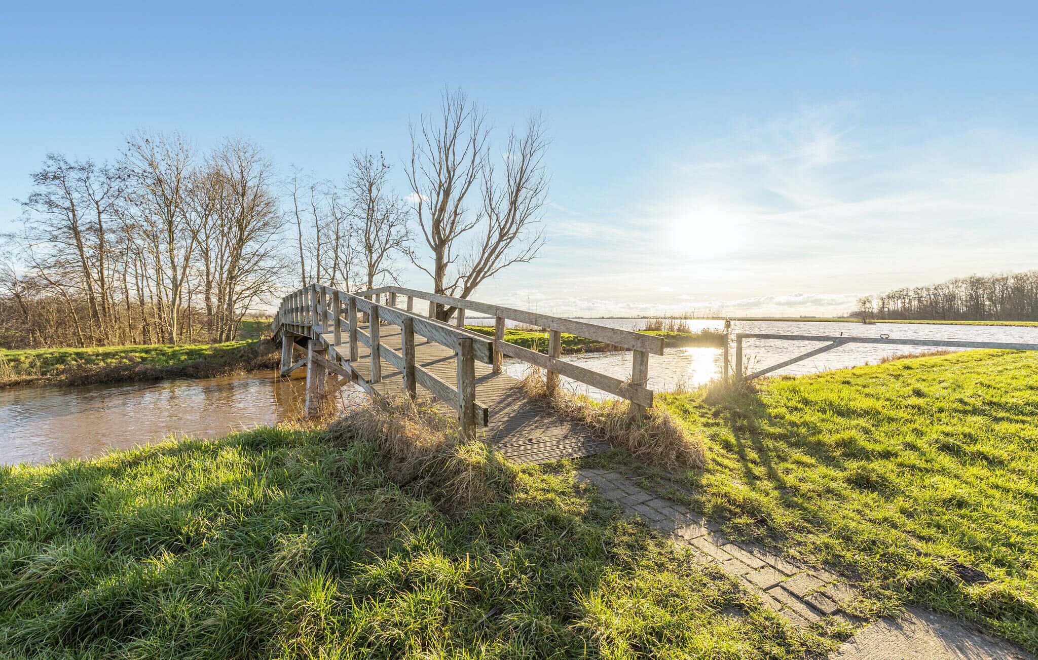 Un pont en bois enjambant une rivière paisible, entouré de berges verdoyantes et d’arbres sans feuilles.