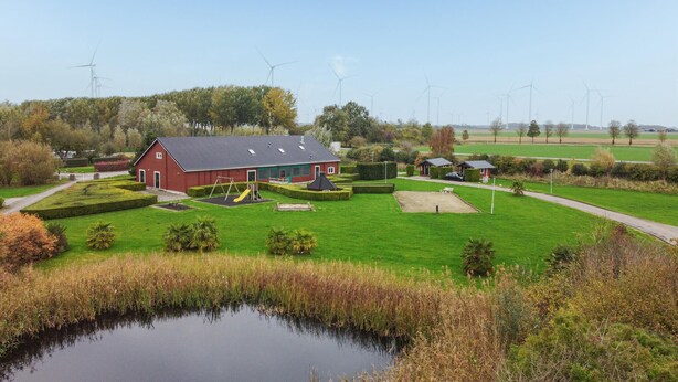 Een rode schuur met een speeltuin, omringd door groene velden en windturbines op de achtergrond.