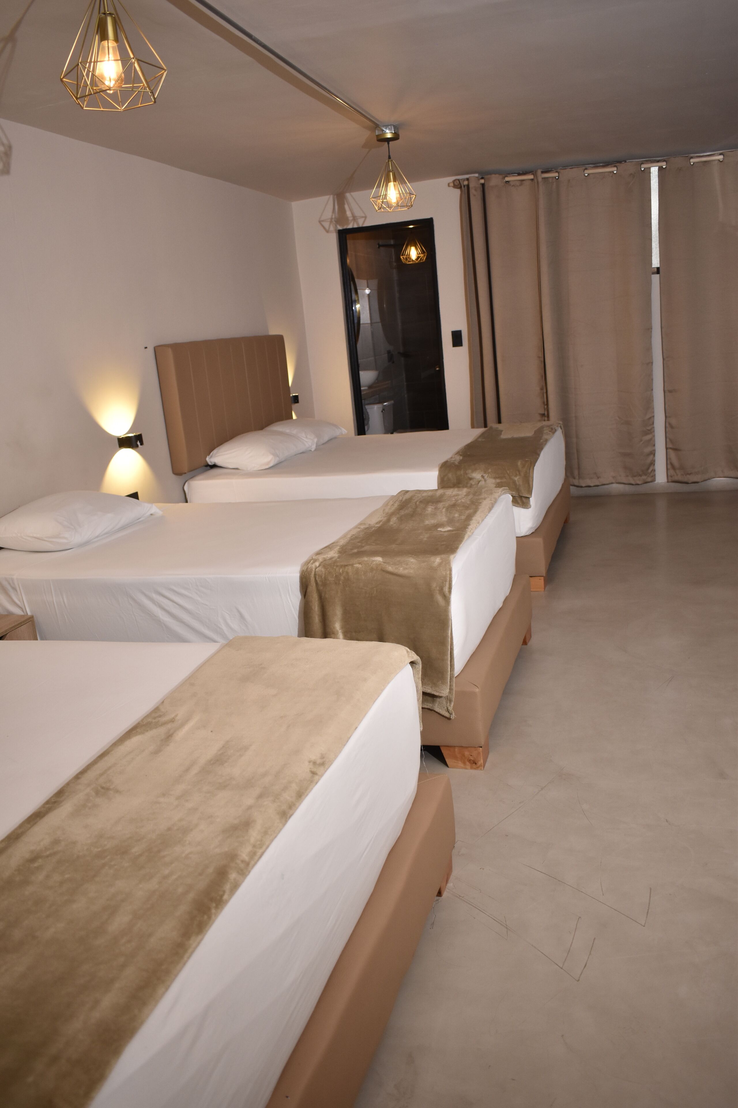 Habitación de hotel con tres camas, cada una con cabecero de madera y ropa de cama blanca. Hay dos luces fijadas a la pared y una puerta con espejo.