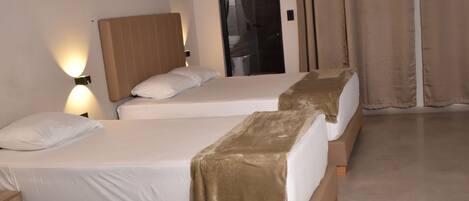 Habitación de hotel con tres camas, cada una con cabecero de madera y ropa de cama blanca. Hay dos luces fijadas a la pared y una puerta con espejo.