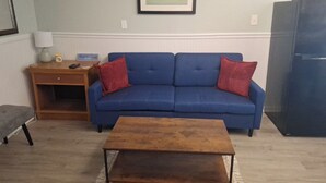 Een woonkamer met een blauwe bank, een houten salontafel, een klein houten bijzettafeltje met een lamp, en een lijst met een foto aan de muur.