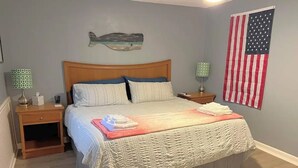 Een netjes opgemaakt bed met een witte quilt, twee kussens en een opgevouwen plaid, tegen een houten hoofdbord. Twee nachtkastjes met lampen, een Amerikaanse vlag aan de muur en een kunstwerk op de wand.