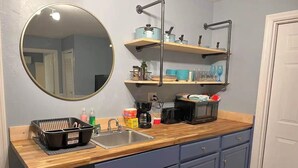 Een keuken met een houten aanrecht, een spoelbak en kastjes. Daarboven bevinden zich planken met diverse keukenbenodigdheden.