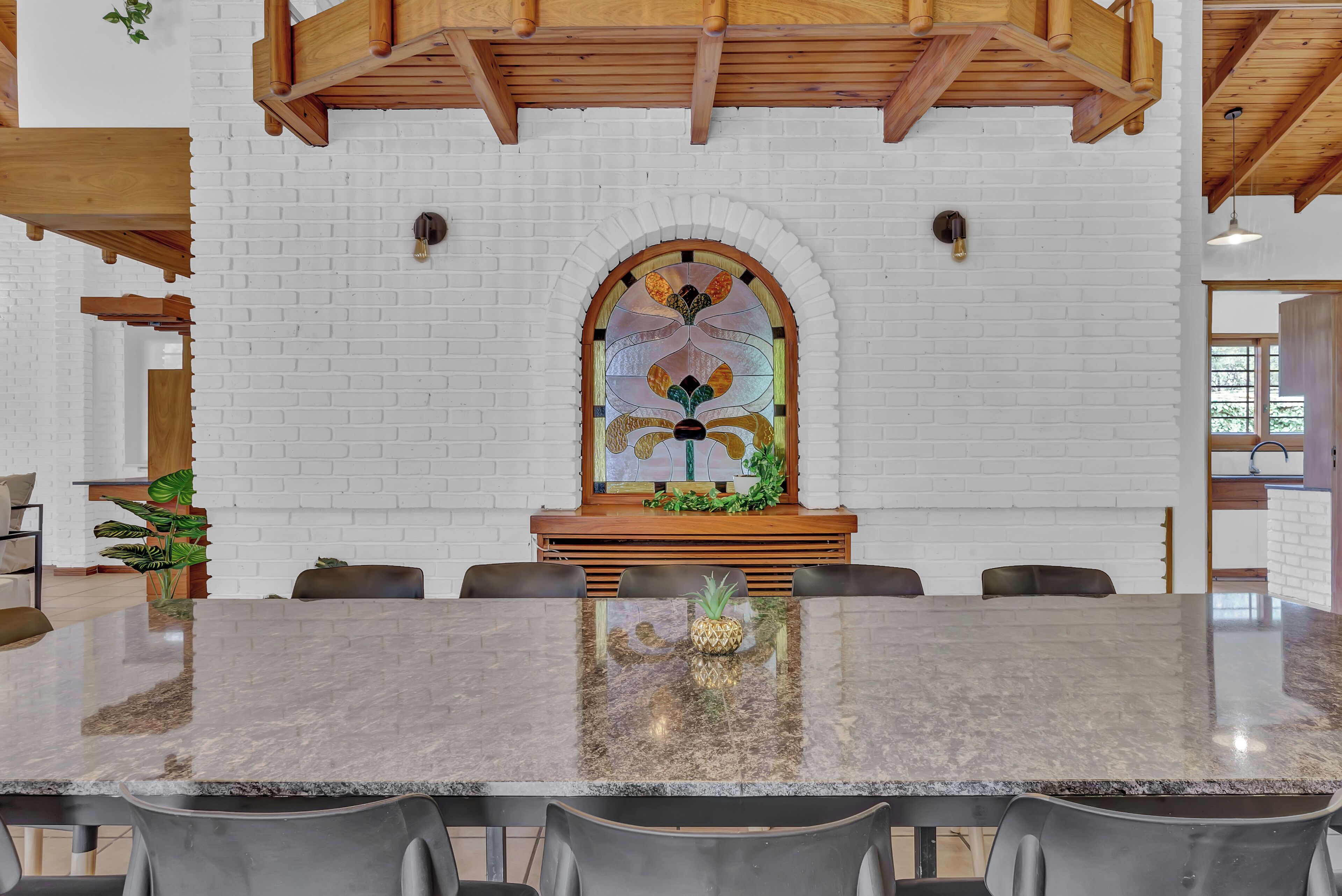 Un comedor moderno con una mesa de mármol, vigas de techo de madera y una pared de ladrillo blanco con un ventanal de vidrio emplomado.