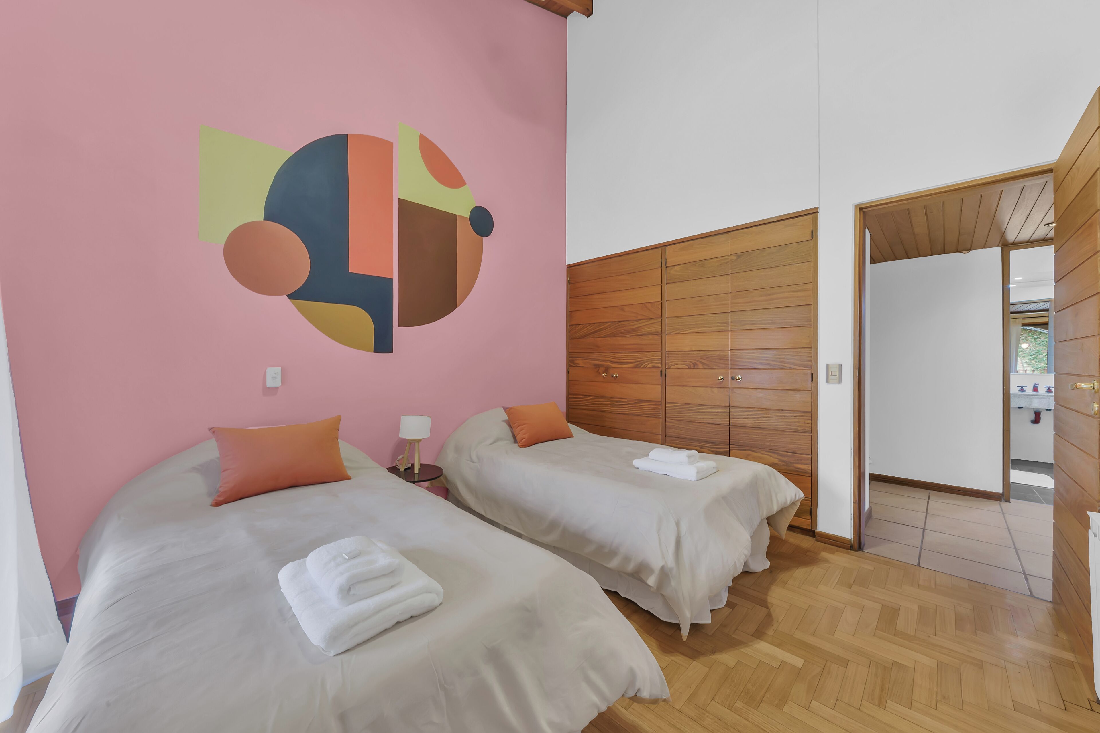 A hotel room with two beds, a wooden wardrobe, a pink wall with abstract art, and a wooden floor.