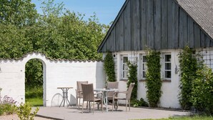 A rustic outdoor seating area with wicker chairs and a table, adjacent to a white building with a dark roof and ivy-covered walls. - Nice home in Farsø with WiFi (Farsø)