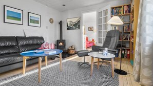 A cozy living room with a black leather sofa, a blue coffee table, a grey armchair, and a bookshelf filled with books. A fireplace with a chimney is visible in the background. - Nice home in Farsø with WiFi (Farsø)