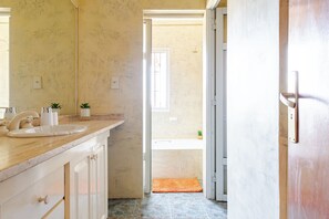 Une salle de bain avec un comptoir en marbre, un lavabo et un miroir. Une porte donne accès à une cabine de douche où se trouve une petite plante sur le comptoir.