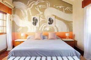 Une chambre avec un grand lit, deux tables de chevet avec des lampes, un mur orné d’œuvres d’art abstraites et une fenêtre avec des rideaux.