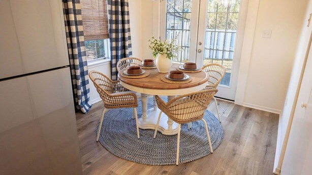 Un coin repas avec une table ronde, des chaises en osier, un vase de fleurs et une vue à travers une porte vitrée.