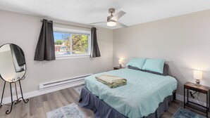 A bedroom with a bed, a ceiling fan, a window with curtains, a bedside table with a lamp, and a mirror. - Cozy home in Logan, close to USU (Logan)